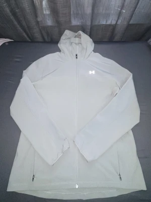 Ljusgrå vindjacka från Under Armour - Snygg ljusgrå vindjacka från Under Armour med huva och hel dragkedja. Jackan har två fickor med dragkedja och reflekterande logga både fram och bak. Perfekt för blåsiga dagar och sportiga outfits.