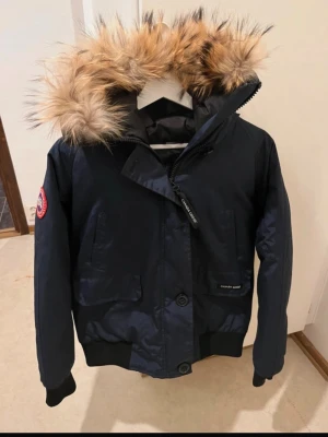 Mörkblå parkas från Canada Goose - Snygg mörkblå parkas från Canada Goose med stor huva och äkta pälsdetalj. Jackan har två stora fickor framtill, ribbade muddar och klassisk logga på ärmen. Perfekt för kalla dagar och riktigt varm. Stängs med dragkedja och knappar.