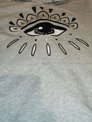 Grå Kenzo hoodie med ögonmotiv - Snygg grå hoodie från Kenzo med det ikoniska ögonmotivet i svart, vitt och blått på bröstet. Tröjan har huva och magficka, samt ribbade muddar vid ärmslut och nederkant. Perfekt för dig som gillar streetwear och vill sticka ut.