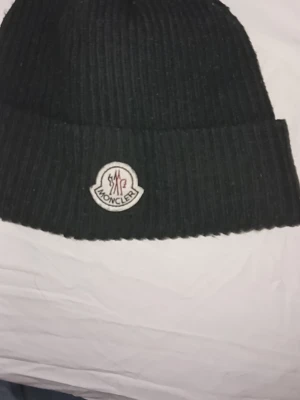 Svart Moncler mössa i ull - Säljer en svart ribbstickad mössa från Moncler med klassisk uppvikt kant och Moncler-logga framtill. Mössan är tillverkad i mjuk ull och passar perfekt för kalla dagar. Enkel och stilren design som funkar till det mesta.