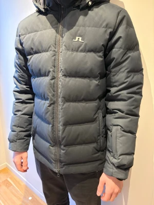 Mörkblå J.Lindeberg vinterjacka storlek M - Snygg mörkblå pufferjacka från J.Lindeberg med huva och dragkedja framtill. Jackan har ett stilrent broderat logotyp på bröstet och breda, vadderade paneler som ger en modern look. Perfekt för kalla dagar och riktigt varm.