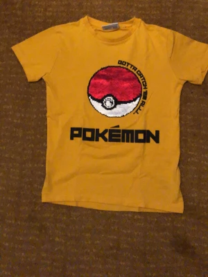 Gul Pokémon T-shirt med Pokéboll - Cool gul T-shirt med stort tryck av en röd och vit Pokéboll och texten 'Gotta catch 'em all' samt Pokémon-loggan på framsidan. T-shirten har rund hals och korta ärmar. Perfekt för dig som älskar Pokémon och vill sticka ut med en färgglad stil.