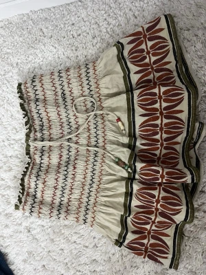 Bohemisk mönstrad kjol med snörning - Säljer en bohemisk kortkjol med elastisk midja och snörning med pärlor. Kjolen har ett unikt mönster i beige, grön och brun med dekorativa detaljer och volang nedtill. Perfekt för sommarens alla äventyr!