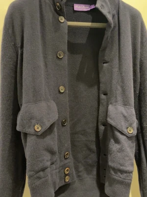 Ralph lauren kofta - Stilren mörkgrå kofta från Ralph Lauren Purple label, tillverkad i Italien. Koftan har ribbstickad struktur, knäppning framtill med flera knappar och två stora fickor med lock och knapp på framsidan. Perfekt för lager på lager och en klassisk look du ser defekterna de är hål lite här och där ungefär 7 hål går att fixa lappen har också lossnat halvt men den är kvar allt går att lösas hos skräddare för någon hundring