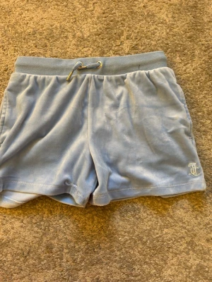 Ljusblå velourshorts Juicy Couture - Mysiga ljusblå shorts från Juicy Couture i mjuk velour med snörning i midjan och broderad JC-logga på benet. Perfekta för chill dagar eller när du vill ha något bekvämt och snyggt.
