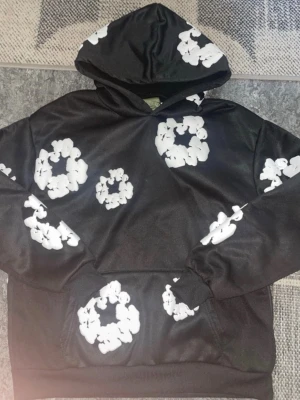Svart hoodie från Denim Tears - Säljer en svart hoodie från Denim Tears med vita blomliknande tryck över hela plagget. Hoodien har en klassisk huva och magficka framtill. Materialet känns mjukt och bekvämt, perfekt för en avslappnad streetwear-look.
