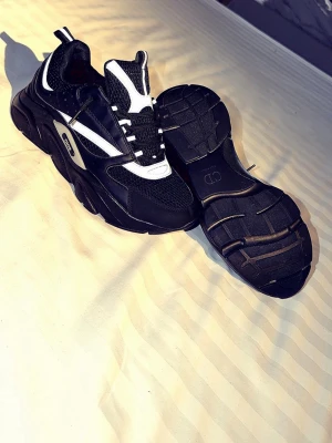 B22 reflect - Chunky sneakers från Dior i svart och vitt med mesh och mockadetaljer. Tjock sula med tydlig Dior-logga undertill och coola kontrasterande paneler. Skorna har en sportig vibe och snörning framtill. Perfekta för dig som gillar statement sneakers.