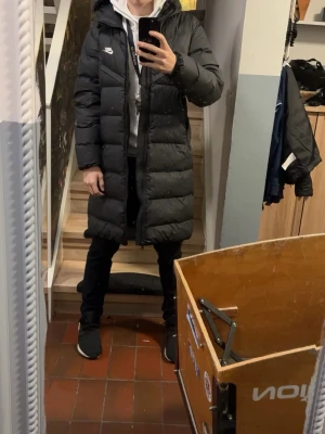 Svart lång dunjacka från Nike - Säljer en svart lång dunjacka från Nike med huva och dragkedja framtill. Jackan har en stilren design med diskret Nike-logga på bröstet och stora, quiltade partier som ger en schysst look. Perfekt för kalla vinterdagar.