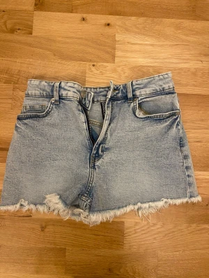 Ljusblå jeansshorts med fransig kant - Säljer ett par ljusblå jeansshorts från H&M med fransig rå kant nertill. Klassisk femficksmodell med bälteshällor och knappgylf. Perfekta för sommaren och ger en avslappnad vibe.
