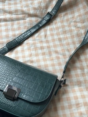 Grön axelväska i croco-mönster - Snygg grön axelväska från Caractère med croco-mönstrad yta och justerbar axelrem. Väskan har ett stilrent silverfärgat spänne framtill och är tillverkad i konstläder. Perfekt accessoar för att lyfta din outfit.