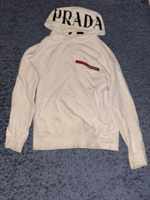 Vit hoodie från Prada med logga - Säljer en vit hoodie från Prada med stor svart logga på huvan och en röd detalj med Prada-tryck på bröstet. Hoodien har klassisk känguruficka och ribbade muddar. Perfekt för dig som vill ha en clean men ändå lyxig look.