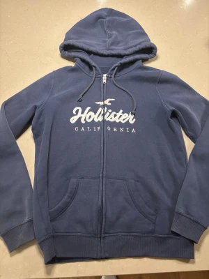 Blå hoodie från Hollister - Säljer en blå zip hoodie från Hollister storlek M. Den är i bra skick och har inga fläckar. Kontakta mig om du vill ha mer info