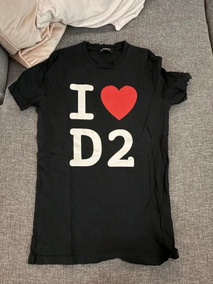 Svart Dsquared2 t-shirt med tryck - Snygg svart t-shirt från Dsquared2 med stort tryck på framsidan där det står 'I ❤️ D2' i vitt och rött. Klassisk rund halsringning och korta ärmar. Perfekt för dig som gillar streetwear och vill sticka ut med ett ikoniskt motiv. Ordinarie pris 2500kr