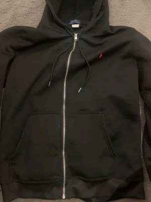 Svart hoodie från Polo Ralph Lauren - Klassisk svart hoodie från Polo Ralph Lauren med dragkedja framtill, två stora fickor och justerbar huva. Liten röd logga broderad på bröstet. Tillverkad i mjukt bomullsmaterial som är skönt att bära. Perfekt för en avslappnad och stilren look.