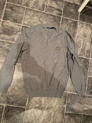 Grå tröja från Lyle & Scott - Grå långärmad tröja från Lyle & Scott med klassisk gul logga på bröstet. Tröjan har v-ringning och ribbade muddar vid ärmslut och nederkant. Perfekt till en avslappnad stil och enkel att matcha.