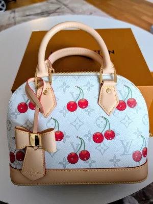 Iouis Vuitton Väska  - Supersöt vit handväsk med körsbärsmönster och beige detaljer. Väskan har en stor rosett framtill, guldfärgade metalldetaljer och röd insida. Perfekt storlek för det viktigaste och har både handtag och axelrem  .                                                                            .Premium Kvalitet ✅ Identisk Design ✅ Hållbara Material✅ Perfekt Finish ✅