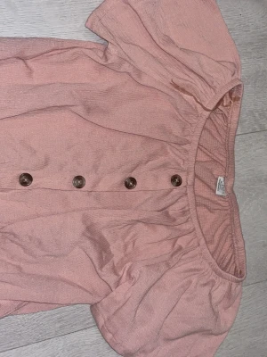 Rosa blus med knappar från Lindex - Söt ljusrosa blus från Lindex med korta puffärmar och dekorativa bruna knappar framtill. Blusen har en rundad halsringning med resår och är tillverkad i ett mjukt, strukturerat tyg som känns luftigt och bekvämt.