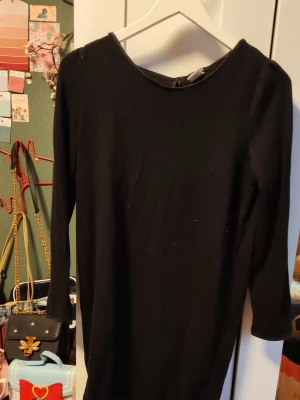 Svart klänning med rosetter i ryggen - En svart, enkel klänning med trekvartsärm och tre söta rosetter i satin baktill. Klänningen har en rak passform och är tillverkad i ett mjukt material som känns bekvämt mot huden. Perfekt för dig som gillar stilrena detaljer och en klassisk look.