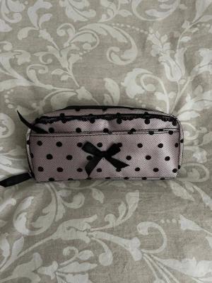 Rosa necessär med svarta prickar och rosett - Söt necessär i ljusrosa tyg med svarta prickar och dekorativ svart rosett framtill. Väskan har dubbla dragkedjor och är täckt med ett tunt svart nät. Perfekt för smink eller småprylar.