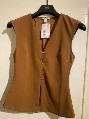 Brun väst med knappar och v-ringning - Snygg brun väst från H&M med djup v-ringning och dekorativa guldfärgade knappar framtill. Västen har en figurnära passform och är tillverkad i ett slätt tyg som ger en stilren look. Perfekt att styla över en skjorta eller t-shirt.