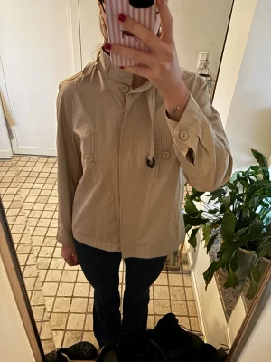 Beige jacka med ståkrage och snörning - Snygg beige jacka med ståkrage och cool snörningsdetalj vid halsen. Jackan har knappar vid ärmslut och två fickor framtill med knappstängning. Perfekt för lager-på-lager och enkel att matcha till olika outfits., midjeband kommer med!!