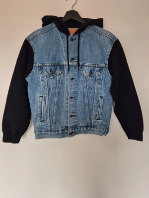 Levi's jeansjacka hybrid med huva - Snygg jeansjacka från Levi's med svarta ärmar och huva i sweatshirtmaterial. Klassisk blå denim med knappstängning, två bröstfickor med lock och två sidofickor. Perfekt för en avslappnad streetstyle-look.