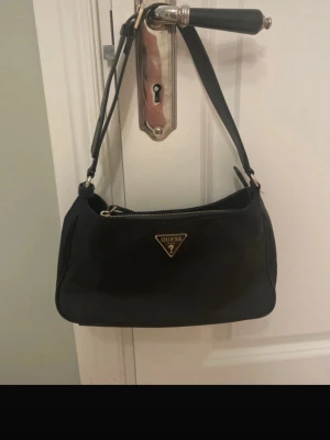 Svart axelväska från Guess - Snygg svart axelväska från Guess med klassisk trekantslogga i guld framtill. Väskan har en enkel, stilren design med dragkedja upptill och justerbar axelrem. Tillverkad i ett slätt material som ger en modern look. 