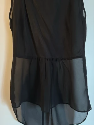 Svart ärmlös festblus från Lindex - Stilren svart blus i ett tunt, transparent material med flowy passform och markerad midja. Blusen är ärmlös och har en längre bakdel för en trendig look. Perfekt att matcha med jeans eller kjol för en snygg outfit.