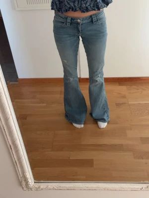 Blå bootcut jeans med broderade fickor - Säljer ett par blå bootcut jeans med slitningar på benen och snygga broderade detaljer på bakfickorna. Jeansen har låg midja och klassisk femficksdesign. Perfekta för dig som gillar en retro vibe och vill sticka ut lite extra.