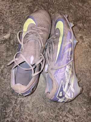 Nike Mercurial lila fotbollsskor - Säljer ett par Nike Mercurial fotbollsskor i lila med stora gula Swoosh-loggor. Skorna har snörning, låg profil och mönstrad ovandel i syntetmaterial för bra bollkontroll. Sulan har fasta dobbar för spel på gräs. Perfekta för dig som vill sticka ut på planen.