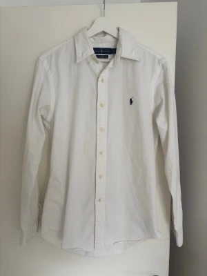 Vit skjorta från Ralph Lauren - Klassisk vit skjorta från Ralph Lauren med den ikoniska broderade loggan på bröstet. Skjortan har lång ärm, knappar framtill och en tidlös krage. Perfekt för en clean och stilren look.