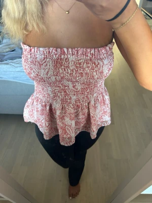 Rödvit axelbandslös bandeautopp - Säljer en söt bandeautopp i rött och vitt med smockad överdel och volang nedtill. Toppen är axelbandslös och har ett blommigt mönster som ger en romantisk vibe. Perfekt till sommaren och lätt att matcha med jeans eller kjol.