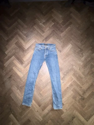 Ljusblå jeans från Nudie - Säljer ett par ljusblå jeans från Nudie Jeans med klassisk femficksdesign och dragkedjegylf. Jeansen har en smal passform och är tillverkade i mjuk denim med subtila slitningar. Perfekta för en avslappnad och trendig look. Size: Midjan är 27 och längden är 30. Mer jeans finns pä min profil