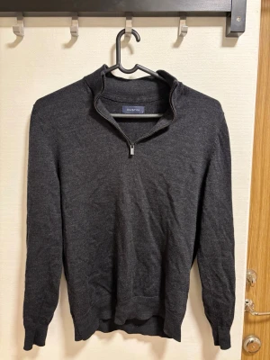 Mörkgrå halvzip-tröja  - Mörkgrå långärmad tröja från Dustin med halvzip och ståkrage. Tillverkad i ett mjukt, tunt stickat material som ger en clean och stilren look. Perfekt för lager-på-lager och funkar året runt.