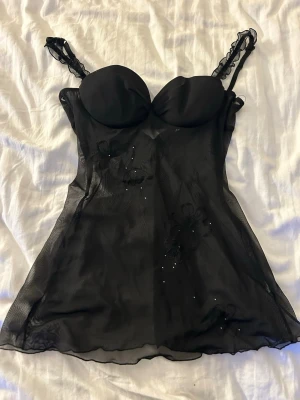 Svart mesh blomstermönstrad linnetopp - Säljer en svart mesh linnetopp med tunna axelband och vadderade kupor. Toppen har blommiga broderier och små glittriga detaljer framtill. Materialet är genomskinligt och ger en lätt och luftig känsla. Perfekt för dig som vill ha något extra till outfiten.