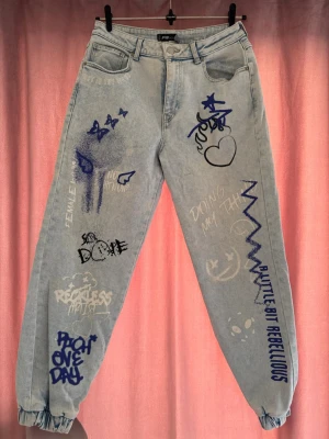 Ljusblå jeans med graffiti-tryck - Unika ljusblå jeans från Pull&Bear med blå och vita graffiti-inspirerade tryck, text och illustrationer över hela benen. Modellen har avslappnad passform, hög midja och resår i bensluten. Perfekta för dig som vill sticka ut med en kreativ streetstyle-look.