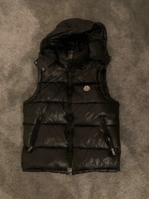 Svart dunväst från Moncler - Snygg svart dunväst från Moncler med avtagbar huva och två dragkedjeförsedda fickor. Västen har Moncler-logga på bröstet och quiltad design som ger en puffig look. Perfekt för lager-på-lager och streetstyle. Inga defekter eller fläckar