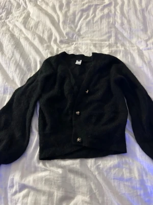 Svart fluffig kofta med knappar - Mysig svart kofta med v-ringning och tre dekorativa knappar framtill. Koftan har en fluffig textur och ballongärmar som ger en trendig siluett. Perfekt att slänga över en topp för en avslappnad men stilsäker look.