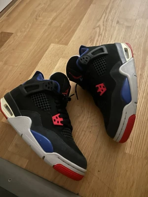 Air Jordan 4 Retro svart/blå/röd - Säljer ett par Air Jordan 4 Retro sneakers med svart ovandel, blå detaljer och röda accenter. Skorna har meshpaneler, plastvingar och Jumpman-logga på tungan. Sulan är vit med blå och röda inslag. Perfekt för dig som vill sticka ut med klassiska Jordans.