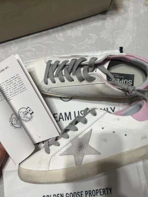 Golden Goose vita sneakers med stjärna - Golden Goose sneakers i vitt läder med grå stjärndetalj på sidan, grå skosnören och ljusrosa häl. Klassisk låg modell med slitna detaljer för en cool vintage-look. Kommer med originalkartong och dustbag.