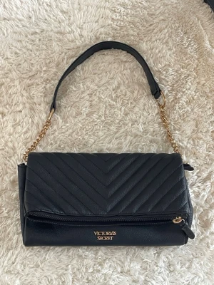 Svart handväska från Victoria's Secret - Stilren svart handväska i konstläder från Victoria's Secret med guldfärgade kedjedetaljer och dragkedja framtill. Väskan har quiltat mönster på locket och magnetlås. Går att använda som axelremsväska eller crossbody. Slitningar på insidan av väskan (se sista bilden) 