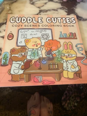 Cuddle Cuties målarbok - En söt målarbok med titeln 'Cuddle Cuties Cozy Scenes Coloring Book'. Omslaget har gulliga djur i labbrockar och boken innehåller mysiga och kreativa scener att färglägga. Perfekt för dig som gillar söta illustrationer och avkoppling.