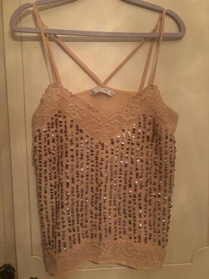 Beige paljettopp från Zara - Supersnygg beige linnetopp från Zara med tunna axelband och korsad rygg. Toppen är dekorerad med glittriga paljetter i vertikala rader och har spetsdetaljer upptill och nedtill. Perfekt för dig som vill sticka ut lite extra.
