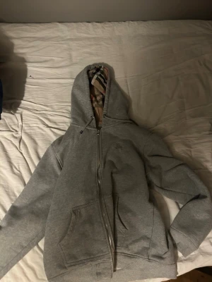 Burberry Hoodie - Säljer en grå hoodie från Burberry med dragkedja framtill och klassiskt rutigt foder i huvan. Hoodien har två stora fickor fram och är gjord i mjukt sweatshirtmaterial. Perfekt för en avslappnad och stilren look. Storlek XL men sitter som L