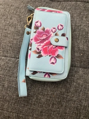 Ljusblå plånbok med blommönster - Snygg ljusblå plånbok med rosa och lila blommor på framsidan. Plånboken har dragkedja runtom, tryckknapp och handledsrem. Insidan har flera kortfack och ett fönster för ID eller foto. Tillverkad i konstläder med mjuk känsla.