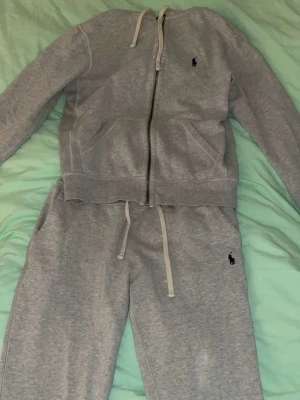 Grått loungewear-set från Polo Ralph Lauren - Mysigt grått loungewear-set från Polo Ralph Lauren med dragkedja på tröjan och matchande byxor med dragsko i midjan. Setet har diskret logga på byxan och är tillverkat i mjukt bomullsmaterial. Perfekt för chill dagar hemma. Priset kan diskuteras 