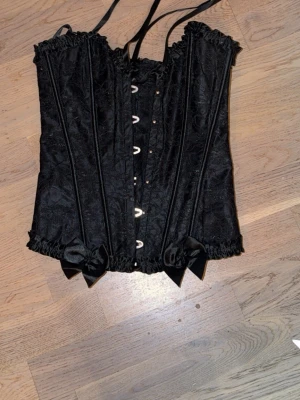 Svart spetskorsett med rosetter - Snygg svart korsett med dekorativa rosetter och spetsdetaljer. Korsetten har snörning bak, hakar fram och fina volangkanter upptill och nedtill. Materialet är glansigt med diskret mönster och ger en elegant siluett. Perfekt för dig som vill ha en klassisk och stilren look.