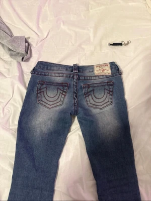 True Religion blå jeans med röd söm - Säljer ett par blå jeans från True Religion med ikoniska röda kontrastsömmar och broderade bakfickor. Klassisk femficksmodell och knappgylf. Jeansen har små skador nere vid fötterna då de var för långa för mig.