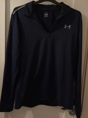 Mörkblå träningströja från Under Armour - Snygg mörkblå långärmad träningströja från Under Armour med halv dragkedja och diskret logga på bröstet. Tillverkad i ett lätt och funktionellt material som passar perfekt för träning. Tröjan har en sportig design med reflexdetaljer på axlarna.
