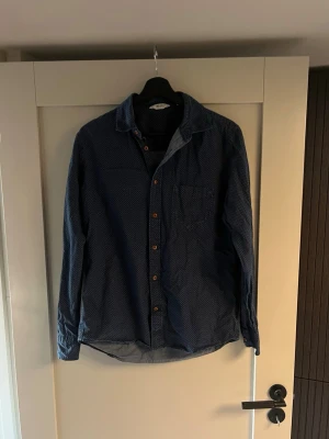 Mörkblå mönstrad skjorta NN07 - Snygg mörkblå skjorta från NN07 med diskret vitt mönster och bruna knappar. Skjortan har klassisk krage, bröstficka och långa ärmar. Perfekt för dig som gillar stilrena detaljer och en schysst look.